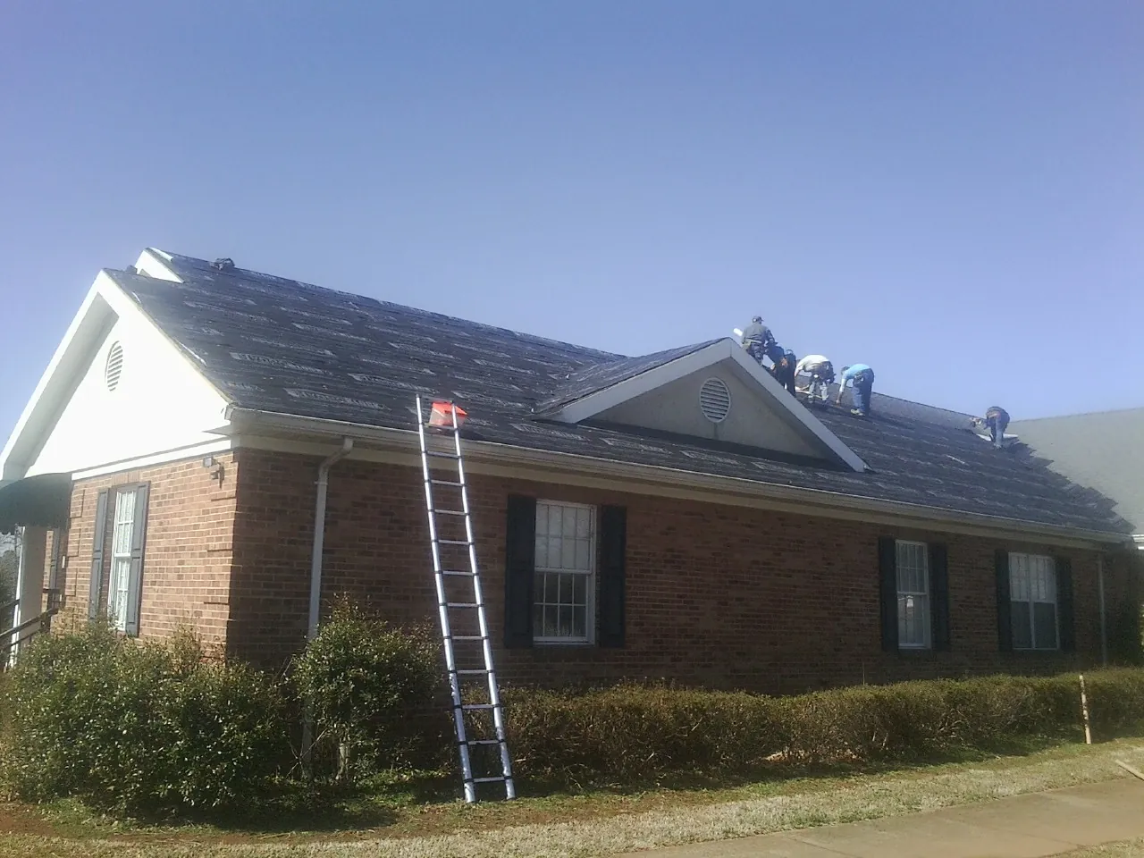 Crew tearing shingles off a brick ranch roof with a ladder set — Metal Roof Repair work by our crew in Chappells