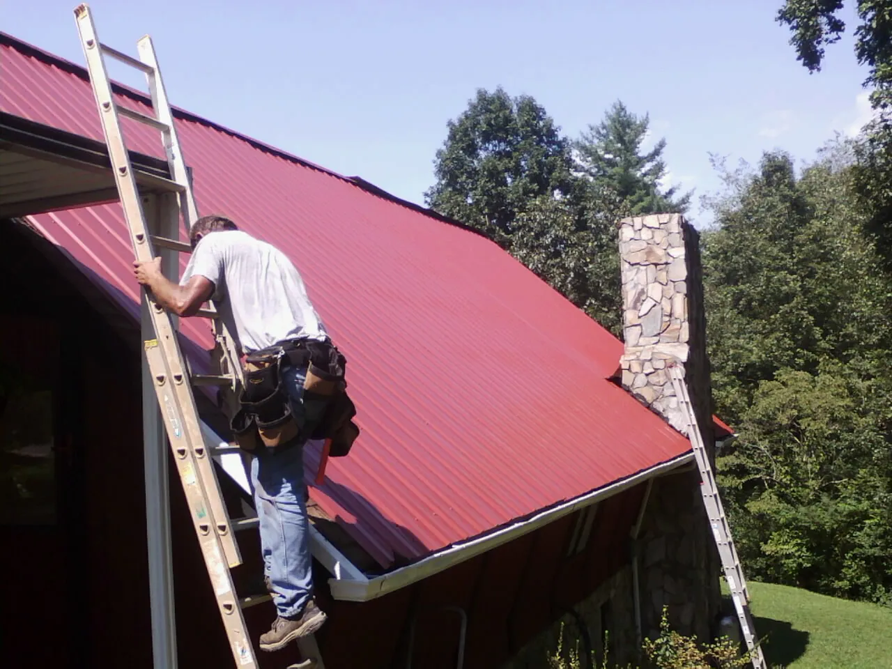 Roofer on a ladder beside a red metal roof and stone chimney — Soffit & Fascia Repair by Roofing Brothers in Chappells